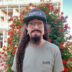 man with glasses, beard, and long dreads in front of red and green plant