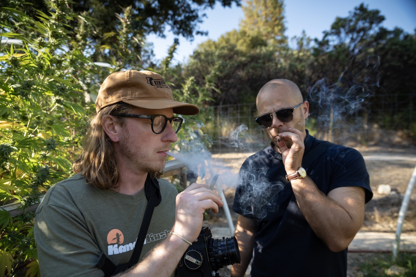 man with long hair smoking a joint on a cannabis farm next to a man with a shaved head smoking a joint on a weed farm