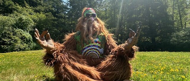 sasquatch sitting cross-legged on grass in front of trees with blue sky