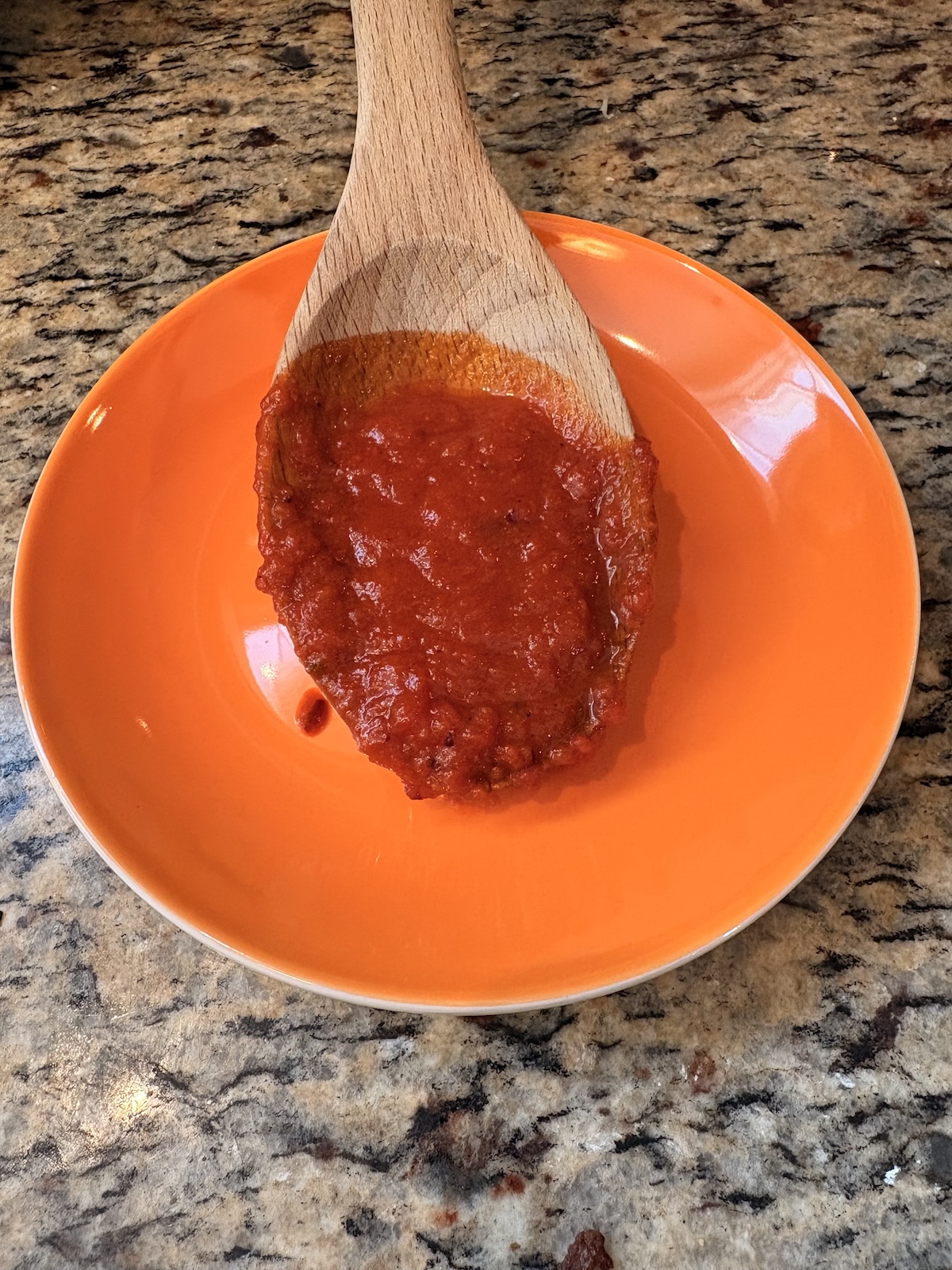 Tomatensoße auf einem Holzlöffel, der auf einem orangefarbenen Teller auf einer Küchenarbeitsplatte liegt